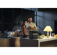 Pár v kuchyni s prenosným generátorom energie, ktorý napája lampu, zatiaľ čo v pozadí beží mikrovlnná rúra.