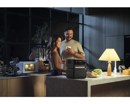 Pár v kuchyni s prenosným generátorom energie, ktorý napája lampu, zatiaľ čo v pozadí beží mikrovlnná rúra.