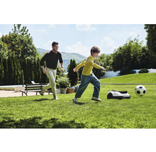 Muž a dieťa hrajú futbal s robotickou kosačkou v záhrade.