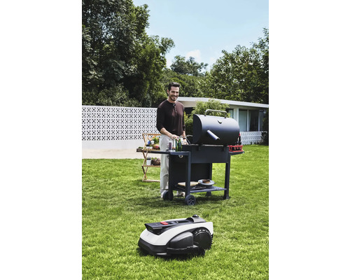 Muž griluje v záhrade s grilom a robotickou kosačkou.