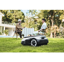 Scéna s robotickou kosačkou na trávu v záhrade, zatiaľ čo dve osoby sedia pri stole