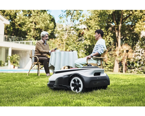 Scéna s robotickou kosačkou na trávu v záhrade, zatiaľ čo dve osoby sedia pri stole