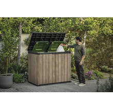 Drevený box na odpadkové koše s dvoma zelenými košmi a osobou, ktorá doň vhadzuje vrece s odpadkami