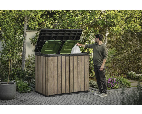 Drevený box na odpadkové koše s dvoma zelenými košmi a osobou, ktorá doň vhadzuje vrece s odpadkami