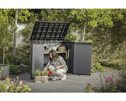 Záhrada so ženou kľačiacou pred otvorenou záhradnou skriňou, v ktorej je uložené záhradné náradie.