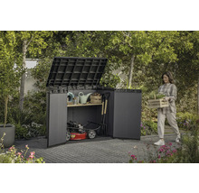 Záhradná scéna so ženou nesúcou debničku s rastlinami vedľa kôlne na náradie plnej záhradného náradia a kosačky na trávu