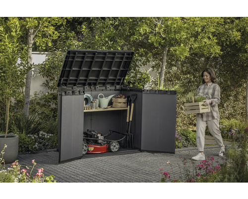 Záhradná scéna so ženou nesúcou debničku s rastlinami vedľa kôlne na náradie plnej záhradného náradia a kosačky na trávu