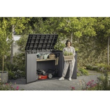 Záhradná scéna s domčekom na náradie, záhradným náradím a ženou držiacou debnu s rastlinami.