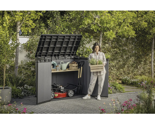 Záhradná scéna s domčekom na náradie, záhradným náradím a ženou držiacou debnu s rastlinami.