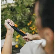 Osoba drží záhradnú hadicu s rozprašovacou hlavicou na pozadí zelených rastlín a žltých kvetov.