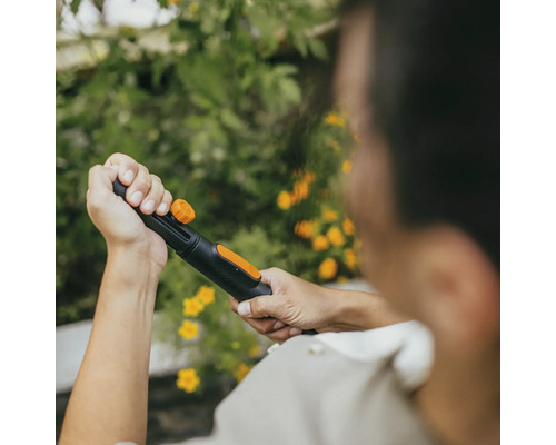 Osoba drží záhradnú hadicu s rozprašovacou hlavicou na pozadí zelených rastlín a žltých kvetov.