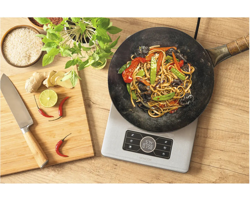 Wok so zeleninou a rezancami na indukčnej varnej doske, vedľa doska na krájanie s nožom, zázvorom, limetkou a chilli