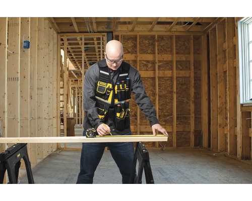 Remeselník meria drevenú dosku pomocou zvinovacieho metra na dvoch kozách v hrubej stavbe.