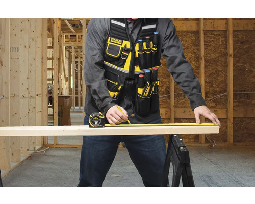 Remeselník meria drevenú dosku pomocou zvinovacieho metra a ceruzkou, má na sebe pracovnú vestu