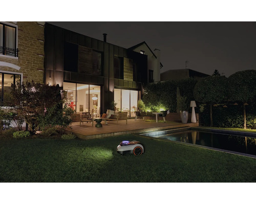 Robotická kosačka na trávu pri nočnej práci v záhrade s domom a bazénom.