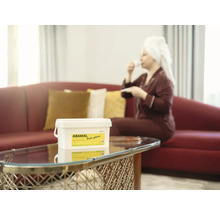 Vedro protiplesňovej farby Abamal Proti Pilisnim na stole, zatiaľ čo žena v pozadí pije kávu.