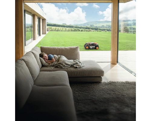 Chlapec spí na gauči, zatiaľ čo robotická kosačka pracuje v záhrade