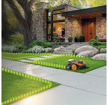 Robotická kosačka na trávniku pred domom s kamennou fasádou a veľkým stromom.