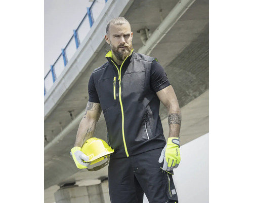 Muž má na sebe pracovnú vestu, tričko, pracovné nohavice, rukavice a drží ochrannú prilbu.