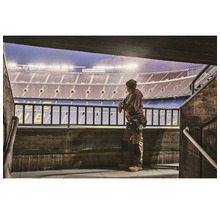 Muž v pracovnom odeve stojí pred štadiónom a pozerá sa na hraciu plochu.