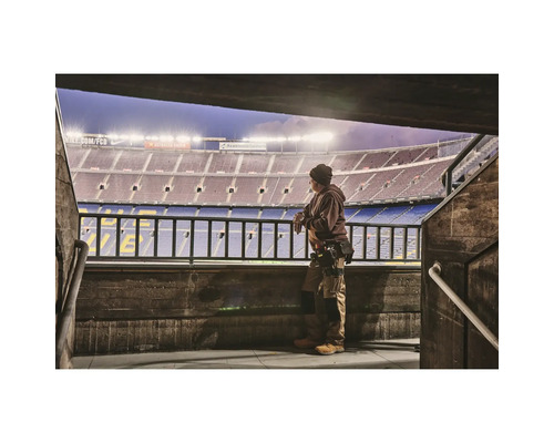 Muž v pracovnom odeve stojí pred štadiónom a pozerá sa na hraciu plochu.