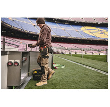 Muž s opaskom na náradie a pracovným odevom pracujúci na elektrickej skrini na štadióne.