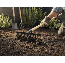 Záhradník hrable zeminu hrabľami, v pozadí je vidieť rýľ a vidly.