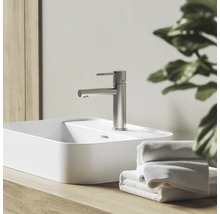 Moderná umývadlová batéria vo vzhľade nehrdzavejúcej ocele na bielom umývadle na dosku, na drevenej doske vedľa uterákov vo svetlej kúpeľni.