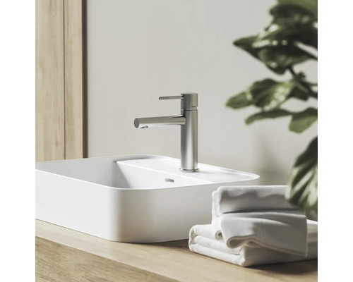 Moderná umývadlová batéria vo vzhľade nehrdzavejúcej ocele na bielom umývadle na dosku, na drevenej doske vedľa uterákov vo svetlej kúpeľni.