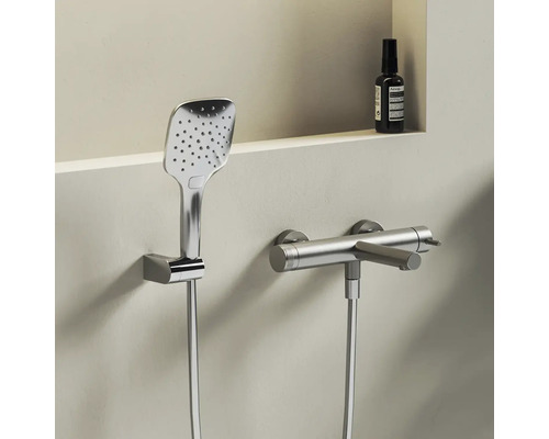 Moderná chrómová sprchová súprava pozostávajúca z hranatej ručnej sprchy v nástennom držiaku a valcovej sprchovej batérie so sprchovou hadicou.
