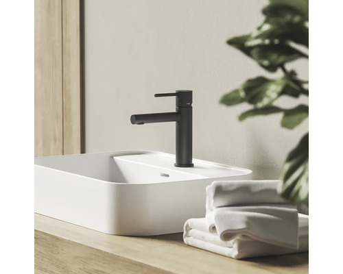 Čierna matná umývadlová batéria na bielom umývadle na dosku na drevenom podklade v kúpeľni s uterákmi.