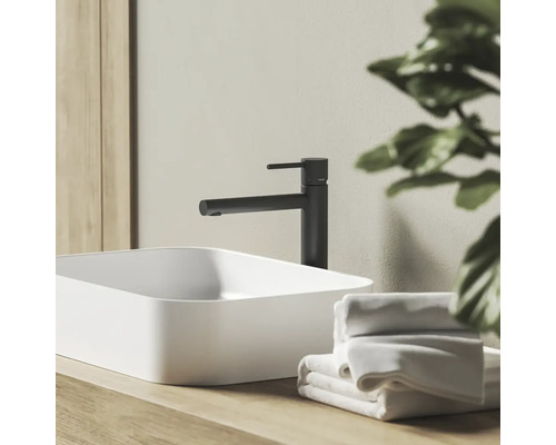 Moderná kúpeľňová scéna s čiernou pákovou umývadlovou batériou a bielym umývadlom na dosku na drevenom podklade s uterákmi.