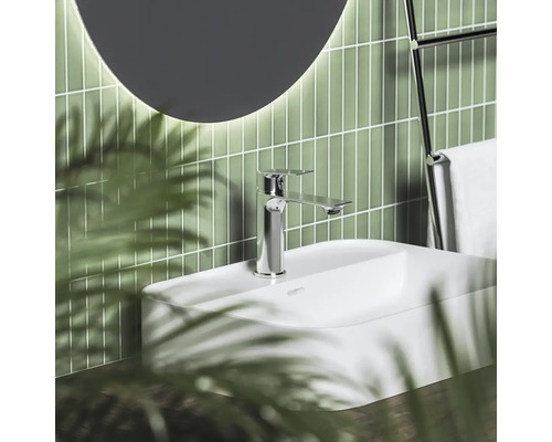 Moderná kúpeľňa s bielym umývadlom, chrómovou batériou, zelenými vertikálnymi obkladmi a okrúhlym zrkadlom.