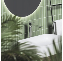 Moderná umývadlová batéria v chróme na bielom umývadle na dosku pred zelenými svislými obkladmi s okrúhlym osvetleným zrkadlom.