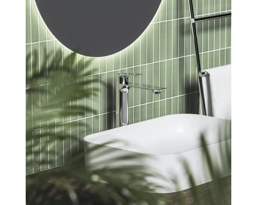 Moderná umývadlová batéria v chróme na bielom umývadle na dosku pred zelenými svislými obkladmi s okrúhlym osvetleným zrkadlom.