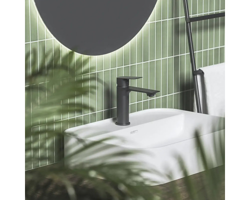 Čierna matná umývadlová batéria na bielom umývadle pred zelenou obloženou stenou s okrúhlym zrkadlom a držiakom na uteráky v modernej kúpeľni.