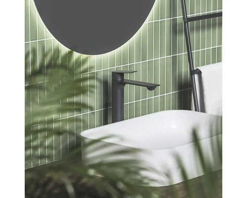 Čierna umývadlová batéria pri bielom umývadle na dosku pred zelenými obkladmi s osvetleným zrkadlom v modernej kúpeľni.