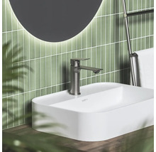 Moderná kúpeľňa s bielym umývadlom na dosku, kovovou batériou, zelenými vertikálnymi obkladmi a okrúhlym podsvieteným zrkadlom.