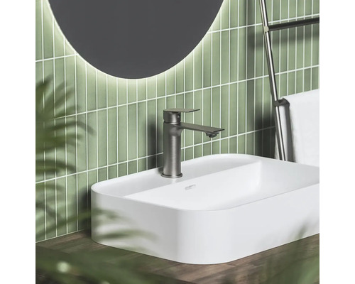 Moderná kúpeľňa s bielym umývadlom na dosku, kovovou batériou, zelenými vertikálnymi obkladmi a okrúhlym podsvieteným zrkadlom.