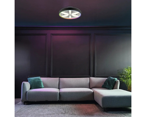 Moderné LED stropné svietidlo s ventilátorom a funkciou farieb RGB, namontované v tmavej obývačke s sivou pohovkou a nástennými panelmi.