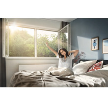 Svetlá spálňa s dokorán otvoreným plastovým oknom, cez ktoré preniká slnečné svetlo, a osobou v posteli, ktorá sa pri prebudení naťahuje.