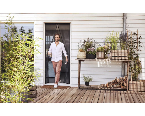 Muž prechádza cez magnetický záves proti hmyzu na terasových dverách na drevenú terasu s mnohými rastlinami v kvetináčoch.