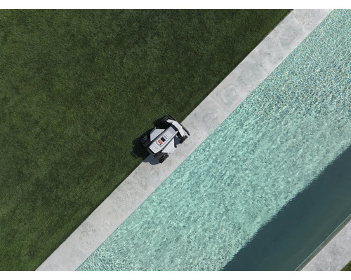 Robotická kosačka na okraji trávnika vedľa bazéna s kamenným lemom, pohľad zhora.