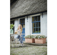 Záhradný kút pred domom so slamennou strechou, bielymi stenami, modrými oknami, veľkými terakotovými kvetináčmi a ženou pri bistro stolíku.