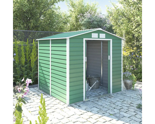 Zelený kovový záhradný domček so sedlovou strechou a bielym rámom, posuvnými dverami a ventiláciou, na kamennej dlažbe vo svetlej záhrade.