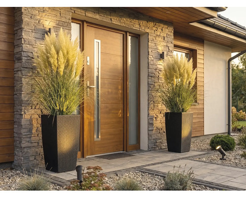 Moderný vchod do domu s drevenými dverami, kamenným obkladom a dvoma vysokými kvetináčmi v ratanovom vzhľade s pampovou trávou.