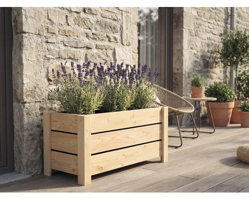 Obdĺžnikový drevený kvetináč s lamelami s kvitnúcou levanduľou na drevenej terase pri kamennom múre.