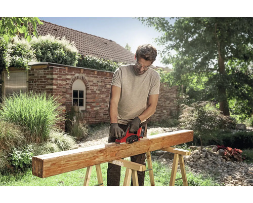 Muž opracováva drevený trám na kozách elektrickým hoblíkom v jasnej záhrade. Má nasadené ochranné okuliare a rukavice.