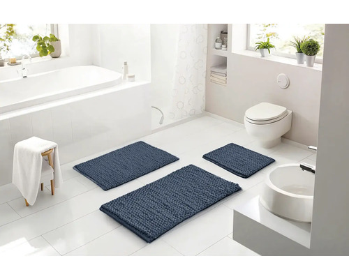 Svetlá moderná kúpeľňa s tromi modrými štruktúrovanými kúpeľňovými predložkami na bielej dlažbe, vrátane vane, toalety a umývadla.