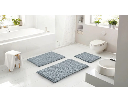 Svetlá kúpeľňa s tromi modrosivými kúpeľňovými predložkami rôznych veľkostí, bielou vaňou, toaletou a umývadlom.